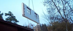 Ein Baustellenbild zeigt ein weißes Gebäudeelement, das von einem Kran in Position gehoben wird. Ein Mann in einem weißen T-Shirt steht auf einer Leiter und beaufsichtigt den Vorgang. Im Hintergrund sind Bäume und ein Holzstapel zu sehen.