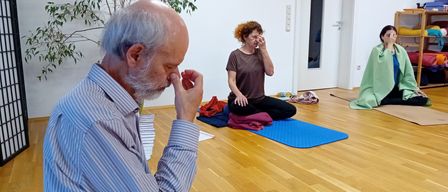Eine Yoga-Sitzung drinnen, bei der ein älterer Mann und zwei Frauen auf Yoga-Matten sitzen. Der Mann meditiert, während die Frauen Yoga-Posen üben. Der Raum hat Holzböden und weiße Wände.