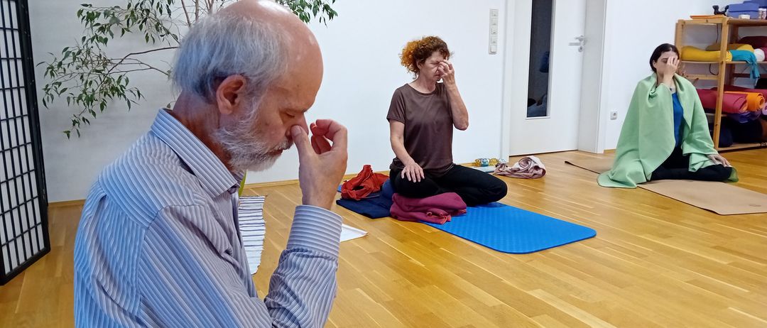 Eine Yoga-Sitzung drinnen, bei der ein älterer Mann und zwei Frauen auf Yoga-Matten sitzen. Der Mann meditiert, während die Frauen Yoga-Posen üben. Der Raum hat Holzböden und weiße Wände.