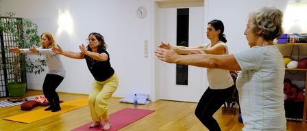 Vier Frauen praktizieren Yoga in einem Raum mit Holzböden. Jede befindet sich auf einer Yogamatte, wobei Matten in verschiedenen Farben sichtbar sind. Ein Topf mit Pflanzen und eine Uhr schmücken die Wand.