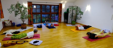 In einem Raum mit Holzboden praktizieren mehrere Personen Yoga. Zwei Personen liegen mit erhobenen Beinen auf dem Rücken, eine andere ruht auf dem Bauch. Yogamatten, ein Topf und ein Faltschirm sind vorhanden.