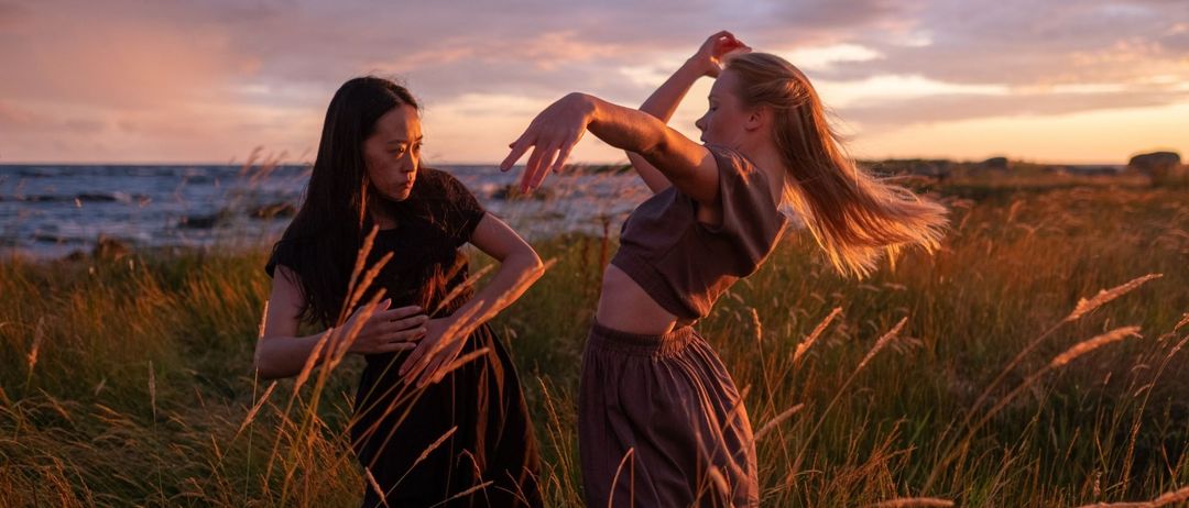 Zwei Frauen tanzen im Freien auf einer Wiese bei Sonnenuntergang. Eine trägt ein schwarzes Kleid, die andere eine braune Bluse.