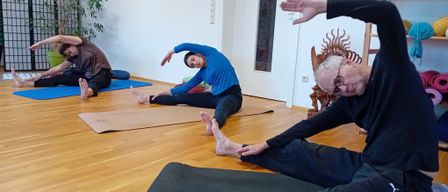 Three people are practicing yoga on mats. One man is sitting cross-legged, another is lying on his side, and a third is lying on his back with arms extended. A clock and plant are in the background.