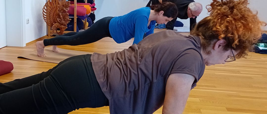 Drei Personen praktizieren Yoga in einem Raum. Eine Frau in einem blauen Shirt und schwarzen Leggings macht Yoga-Posen. Eine weitere Person, ein Mann, macht vor der Frau Yoga-Posen.