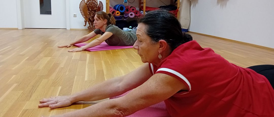 Zwei Frauen praktizieren Yoga auf dem Holzboden. Eine trägt ein grünes Shirt und die andere ein rotes. Im Hintergrund sind verschiedene Yogamatten und Ausrüstungsgegenstände zu sehen.