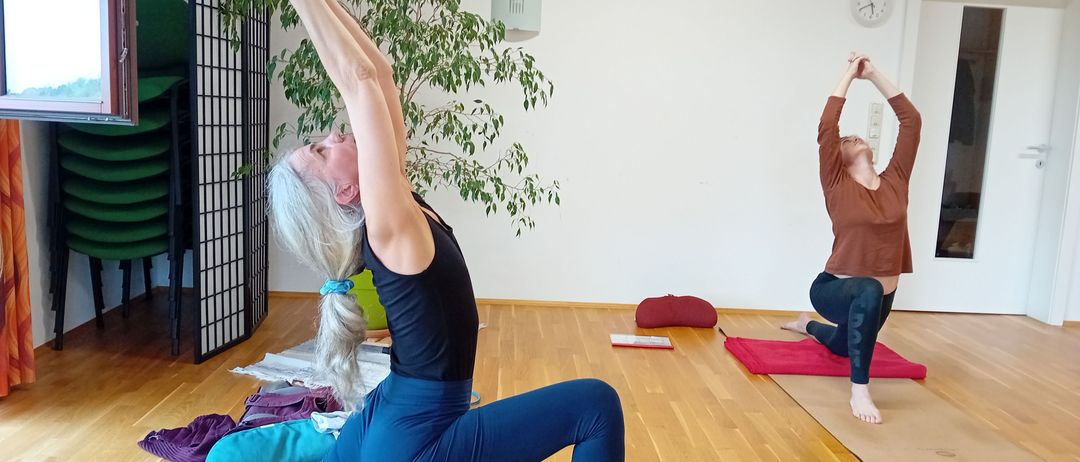 Zwei Frauen praktizieren Yoga in einem Studio. Eine Frau ist in einer Stehposition, während die andere sitzt. Das Studio hat Holzböden, weiße Wände und eine Pflanze.