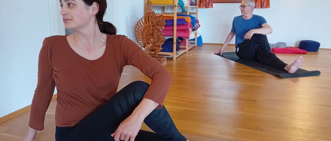 Eine Frau in einem braunen Shirt und schwarzen Leggings sitzt in einer Yoga-Pose auf einer Matte. Hinter ihr sitzt ein Mann in einem blauen Shirt und schwarzen Hosen auf einer Matte, beide in einem Raum mit Holzböden.