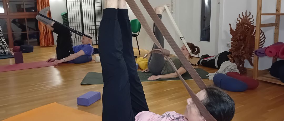 Eine Frau in einem rosa T-Shirt macht Yoga auf einer Matte, liegt auf dem Rücken mit erhobenen Beinen und einem Gurt um die Füße. Andere Menschen machen ebenfalls Yoga im Hintergrund.