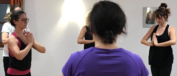 Eine Gruppe von Frauen praktiziert Yoga in einem Raum. Drei Frauen in schwarzen und lila Shirts führen eine Pose aus, während eine Frau in einem weißen Shirt zusieht. Ein Bilderrahmen ist an der Wand.