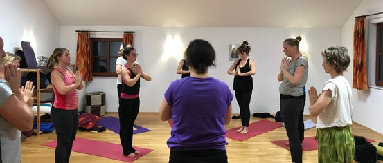 A group of women in yoga attire are practicing yoga in a room with wooden floors. They are standing on yoga mats in various poses.