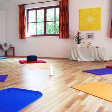 Yogamatten in verschiedenen Farben sind auf einem Holzboden ausgelegt, mit einem in Weiß gedeckten Tisch und einer Topfpflanze in der Nähe. Fenster mit roten Vorhängen und ein großes gelbes Gemälde schmücken den Raum.