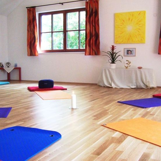 Yogamatten in verschiedenen Farben sind auf einem Holzboden ausgelegt, mit einem in Weiß gedeckten Tisch und einer Topfpflanze in der Nähe. Fenster mit roten Vorhängen und ein großes gelbes Gemälde schmücken den Raum.