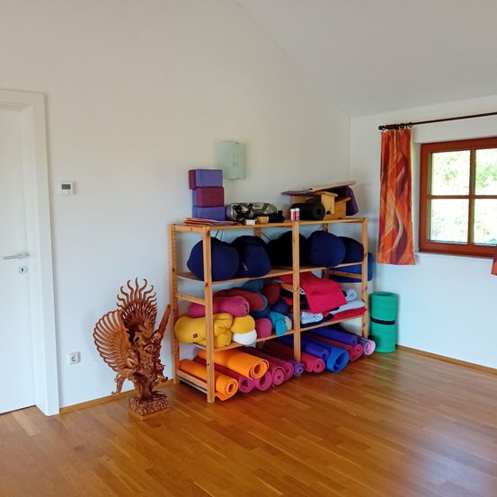 Ein Yoga-Raum mit Holzboden und weißen Wänden. Es gibt ein Holzregal, das mit Yogamatten, Kissen und anderen Yoga-Zubehör gefüllt ist. Ein dekorativer hölzerner Vogel steht auf dem Boden in der Nähe des Regals.