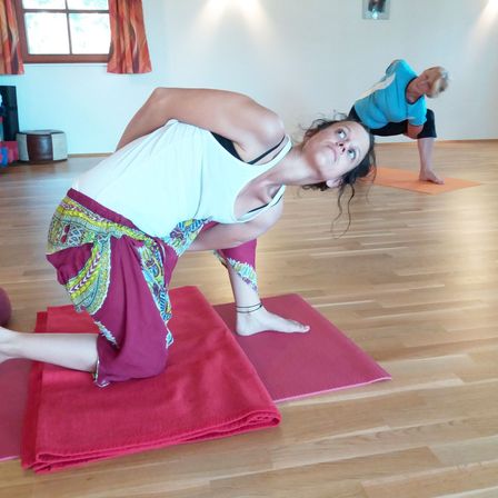 Zwei Frauen praktizieren Yoga in einem Raum mit Holzboden. Eine Frau führt eine Vorbeuge durch, während die andere in einer Plankenposition ist. Es gibt rote Yoga-Matten und orangefarbene Yoga-Blöcke auf dem Boden.