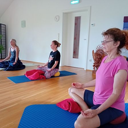 Drei Frauen in einem Raum mit Holzböden machen Yoga. Jede hat eine blaue Matte unter sich. Die Frau im rosa Shirt ist in einer Lotusposition.