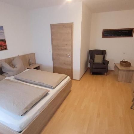 Ein modernes Schlafzimmer mit einem Doppelbett, Holzboden, weißen Wänden und einer Tür. Ein Bild und ein gerahmtes Objekt sind an der Wand. Ein Stuhl und ein Schreibtisch befinden sich rechts.