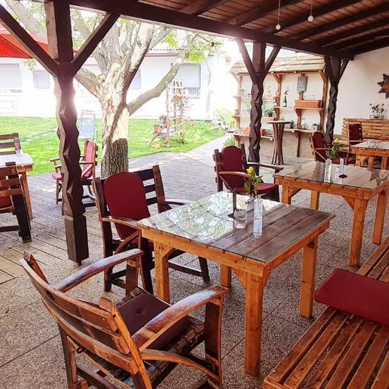 Outdoor-Essbereich aus Holz mit Tischen und Stühlen, einem großen Baum und einem roten Schirm. Jeder Tisch hat eine Blumenvase. Im Hintergrund befindet sich ein Gebäude und ein Rasen.