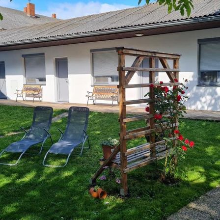 Ein Haus mit grauem Dach und weißen Wänden hat einen Garten mit Bänken, Stühlen und Blumen. Der Rasen ist grün und es gibt Topfpflanzen.