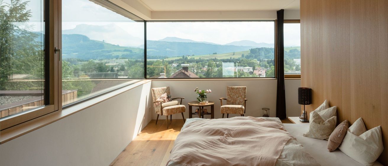 Ein Schlafzimmer mit großen Fenstern, die einen malerischen Blick auf Berge und eine Stadt bieten. Das Bett hat eine rosa Decke, und zwei Stühle mit einem kleinen Tisch halten Blumen.