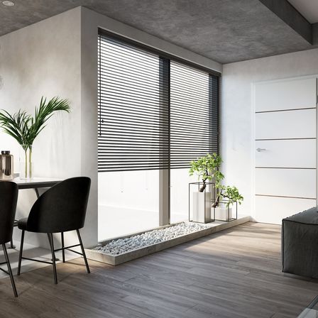 Ein minimalistisches Interieur mit Holzboden, einer weißen Wand, einem schwarzen Stuhl, einem Tisch und einem großen Fenster mit Jalousien. Zwei Topfpflanzen stehen auf der Fensterbank.