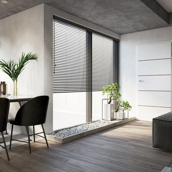 Ein minimalistisches Interieur mit Holzboden, einer weißen Wand, einem schwarzen Stuhl, einem Tisch und einem großen Fenster mit Jalousien. Zwei Topfpflanzen stehen auf der Fensterbank.
