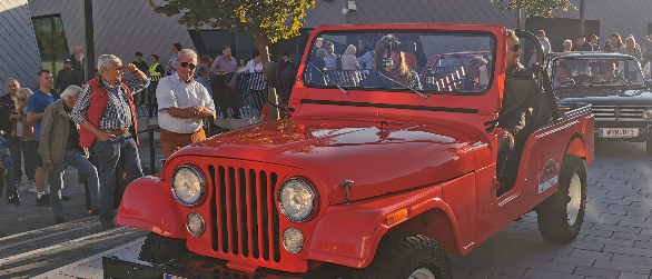 Ein roter Jeep mit offener Tür und einem Mann, der in ein Mikrofon spricht, steht auf der Straße, umringt von Menschen.