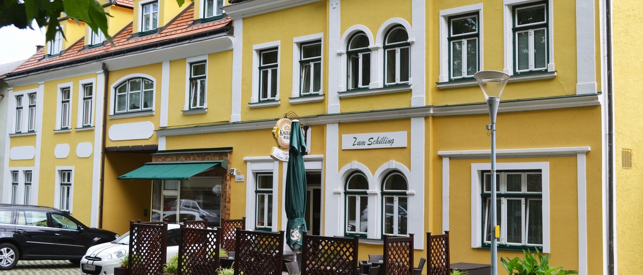 Ein gelbes Gebäude mit weißer Einfassung und rotem Dach hat ein Schild, das 'Gasthof zum Schilling' steht. Es hat bogenförmige Fenster und ein grünes Vordach. Es gibt draußen aufgestellte Tische und Stühle mit einem grünen Schirm. Eine Straßenlampe steht an der Seite.