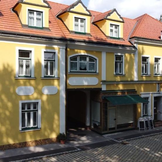 Ein gelbes Gebäude mit weißen Fenstern hat mehrere Fenster und ein rotes Dach. Über dem Eingang befindet sich ein Schild.