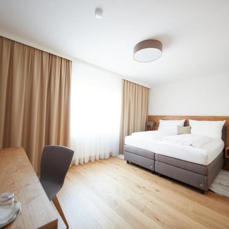 Ein Schlafzimmer mit einem Bett, zwei Kissen und einer Lampe an der Decke. Der Raum hat einen Holzboden und weiße Wände. Ein Schreibtisch mit einem Stuhl steht links.