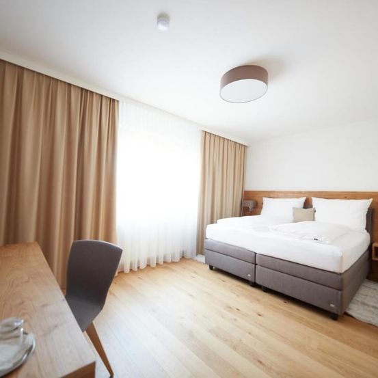 Ein Schlafzimmer mit einem Bett, zwei Kissen und einer Lampe an der Decke. Der Raum hat einen Holzboden und weiße Wände. Ein Schreibtisch mit einem Stuhl steht links.