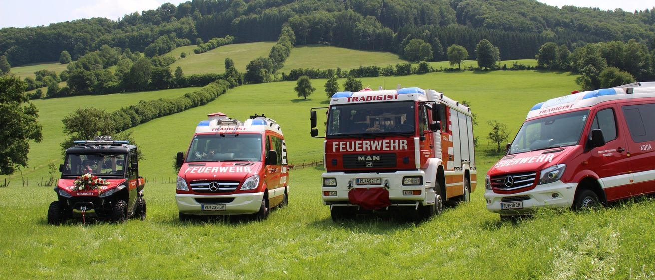 Drei Rettungsfahrzeuge sind auf einer grünen Wiese mit Hügeln im Hintergrund geparkt. Das mittlere Fahrzeug ist ein roter Feuerwehrwagen mit 'Feuerwehr' darauf. Alle Fahrzeuge sind Mercedes-Benz.