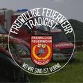 Das Emblem der Freiwillige Feuerwehr Tradigist mit dem Motto 'Wir sind immer da.' Es zeigt einen Feuerwehrhelm, ein rotes Auto und einen Feuerwehrwagen im Hintergrund.