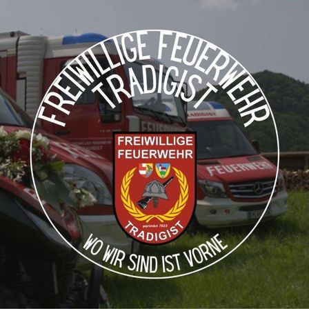 Das Emblem der Freiwillige Feuerwehr Tradigist mit dem Motto 'Wir sind immer da.' Es zeigt einen Feuerwehrhelm, ein rotes Auto und einen Feuerwehrwagen im Hintergrund.