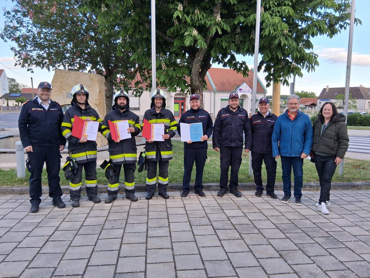 A group of firefighters in uniform holding certificates stand in front of a tree, buildings, and grass.