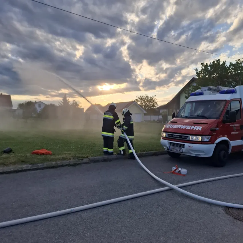 Zwei Feuerwehrleute sprühen Wasser aus einem Schlauch auf der Straße. Ein Feuerwehrwagen ist in der Nähe geparkt. Der Himmel ist bewölkt und die Sonne geht unter.