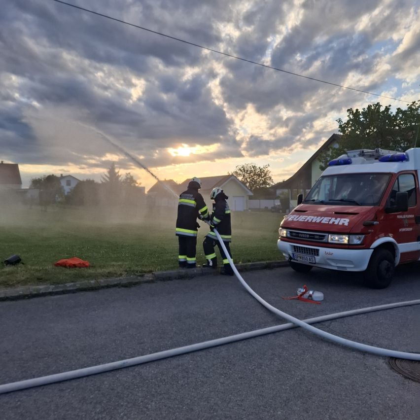 Two firefighters are spraying water from a hose on the road. A fire truck is parked nearby. The sky is cloudy, and the sun is setting.