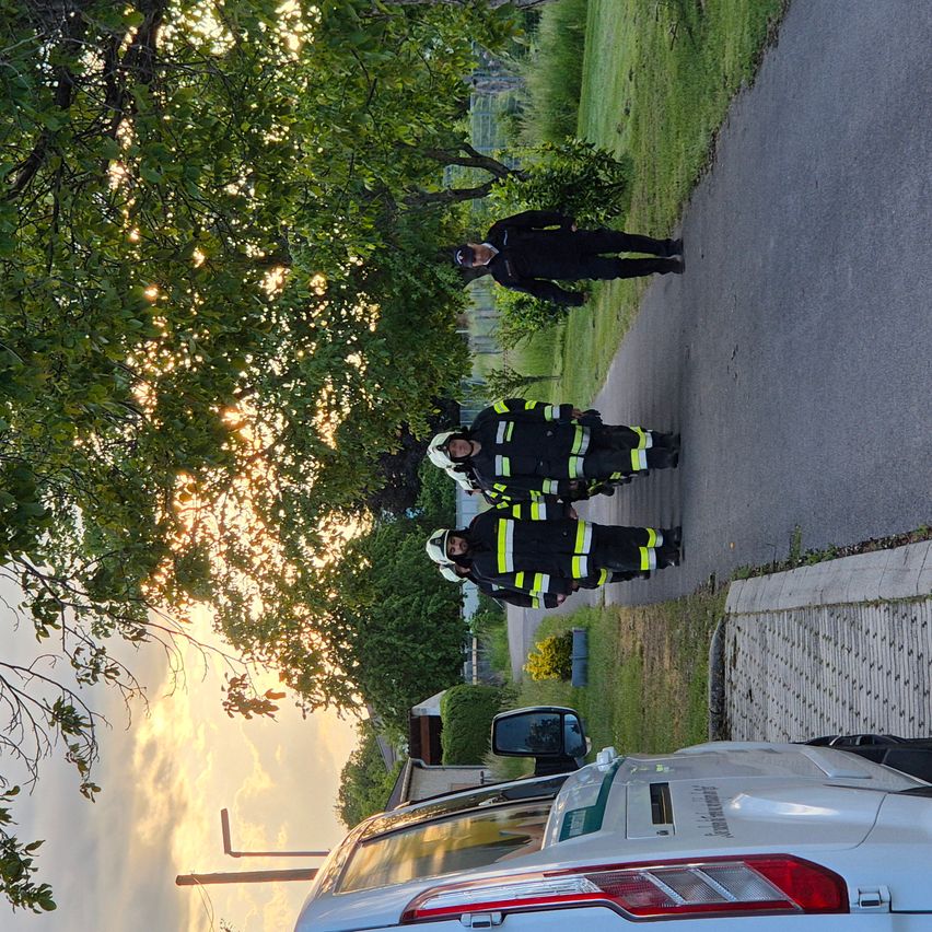 Three firefighters in uniform are standing on a road with a white car parked nearby. The sky is clear with some clouds.