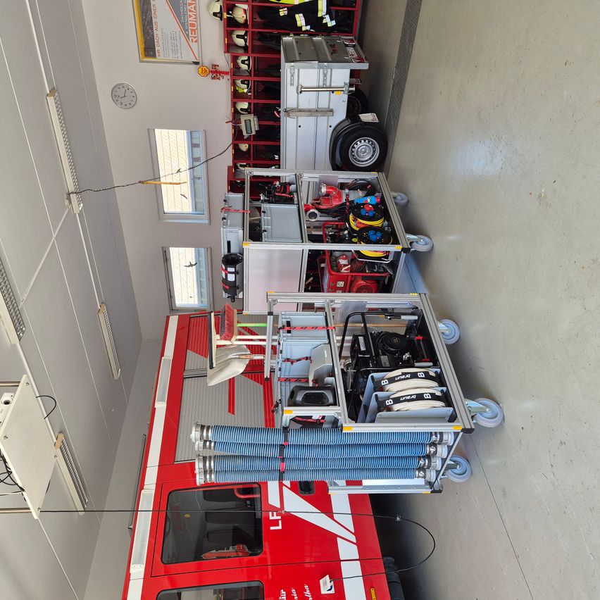 Ein Feuerwehrauto steht in einer Garage geparkt, mit mehreren angehängten Ausrüstungswagen. Die Ausrüstungswagen haben Feuerlöscher, Schläuche und andere Feuerwehrgeräte. Die Garage hat weiße Wände und grauen Boden.