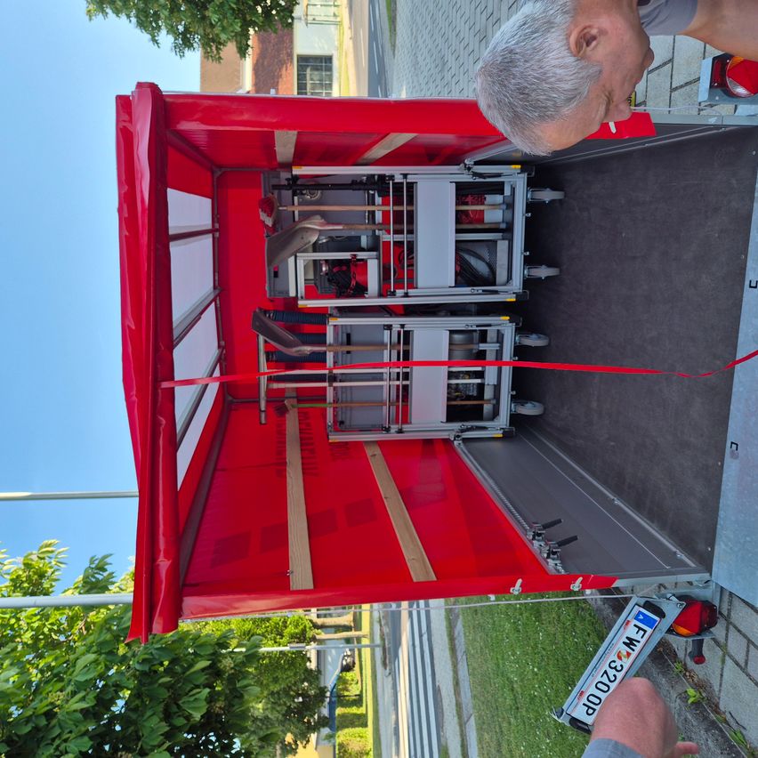 Ein roter LKW mit geöffneter Heckklappe, in dem verschiedene Werkzeuge und Ausrüstung zu sehen sind, einschließlich Schaufeln und anderer Werkzeuge auf einem Metallgestell. Ein Nummernschild ist rechts sichtbar.