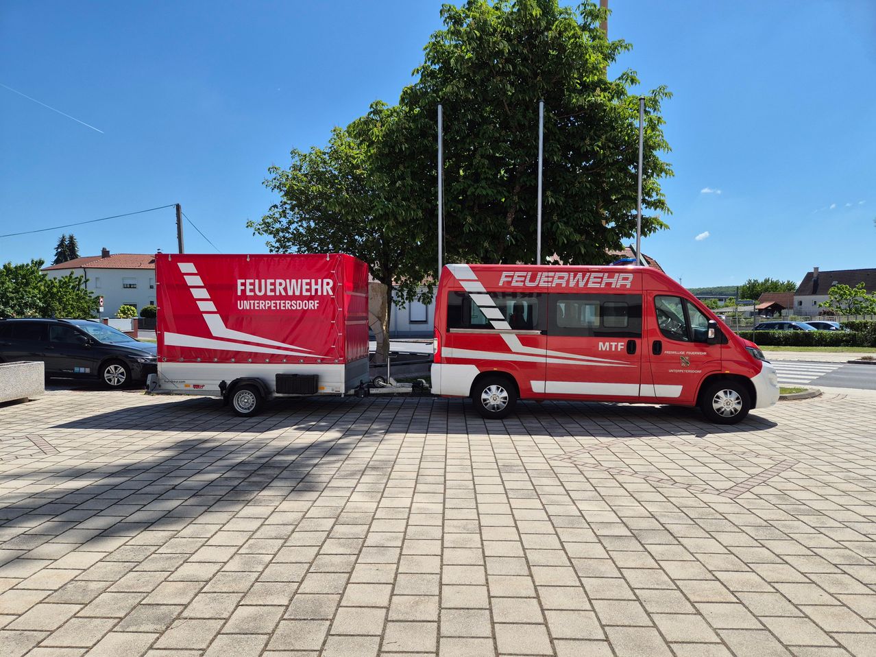 Ein roter Feuerwehrwagen und sein Anhänger sind in einem sonnigen Bereich geparkt, mit einem Baum und einem Haus dahinter. Der Text auf dem Anhänger lautet 'Feuerwehr Unterpetersdorf'.