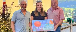 Drei Menschen stehen nebeneinander und halten ein KlimaTicket-Schild. Der Mann auf der linken Seite trägt ein graues Hemd und blaue Shorts. Die Frau in der Mitte hält das Schild und trägt ein schwarzes Shirt. Der Mann auf der rechten Seite trägt ein rosa gestreiftes Hemd.