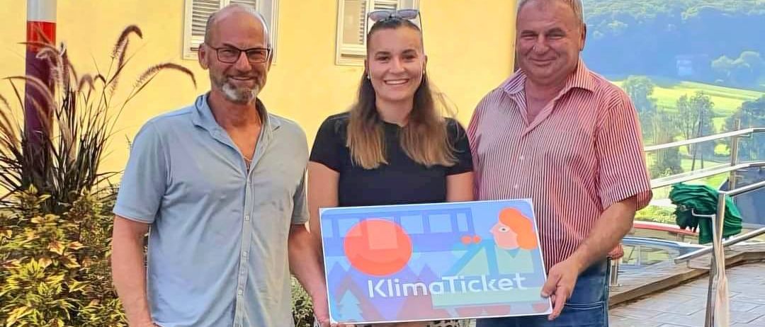 Drei Menschen stehen nebeneinander und halten ein KlimaTicket-Schild. Der Mann auf der linken Seite trägt ein graues Hemd und blaue Shorts. Die Frau in der Mitte hält das Schild und trägt ein schwarzes Shirt. Der Mann auf der rechten Seite trägt ein rosa gestreiftes Hemd.