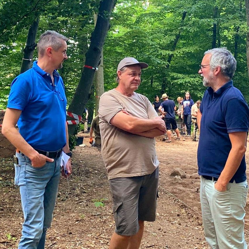 Drei Männer stehen in einer Waldlichtung. Zwei tragen blaue Hemden, einer mit den Händen in den Taschen. Die anderen beiden tragen Hüte, einer mit verschränkten Armen. Hinter ihnen stehen mehrere Menschen und beobachten.