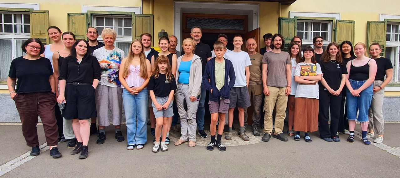 Eine Gruppe von Menschen steht vor einem gelben Gebäude mit mehreren Fenstern und einer offenen Tür und lächelt für ein Foto.
