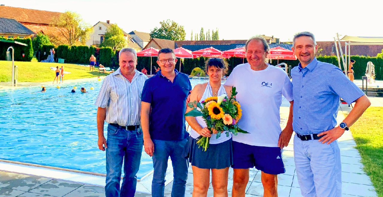 Vier Personen posieren für ein Foto am Schwimmbad, einer hält einen Blumenstrauß. Hinter ihnen sind rote Sonnenschirme und Gebäude.