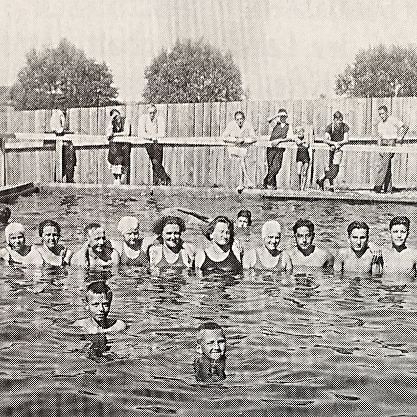 Ein Schwarz-Weiß-Foto zeigt eine Gruppe von Menschen in einem Schwimmbecken, mehrere Erwachsene stehen am Rand und beobachten.