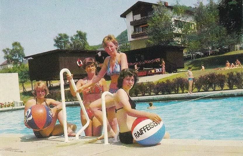 Vier Frauen posieren mit Strandbällen am Rand eines Pools. Im Hintergrund sind Menschen und ein Gebäude mit Balkonen zu sehen.