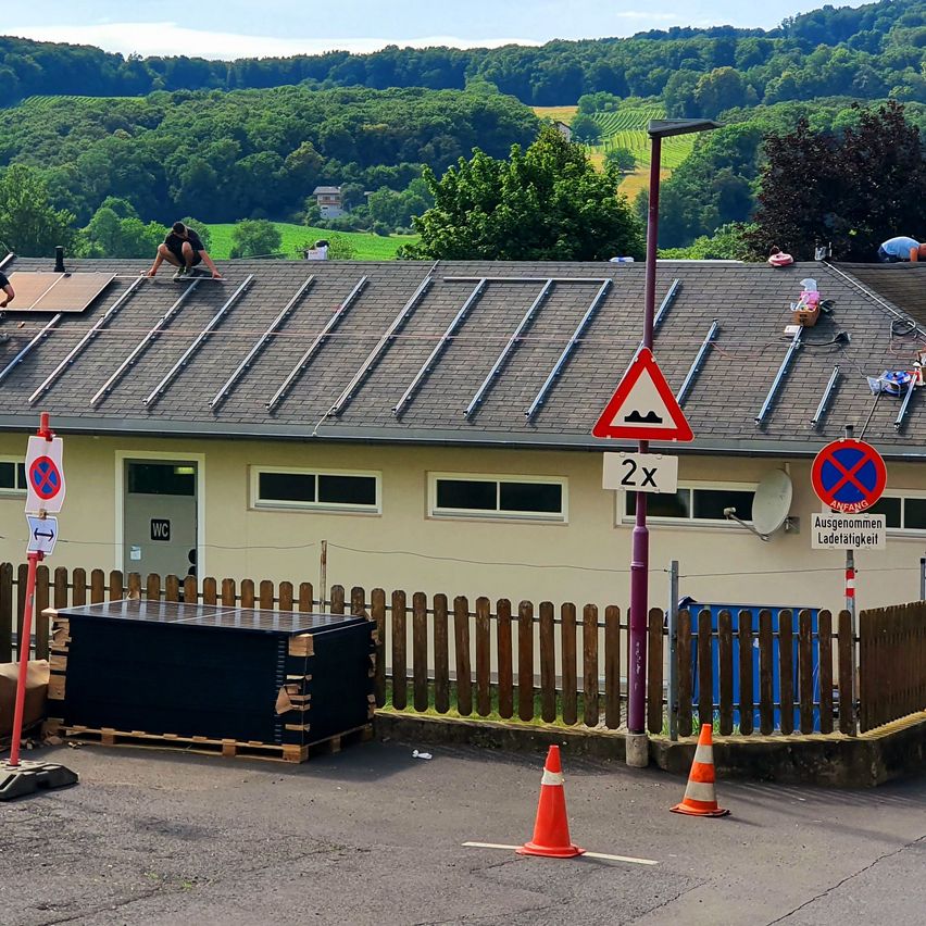 Arbeiter installieren Solarpanels auf dem Dach eines Gebäudes. Das Dach ist mit einem Zaun umgeben, der ein Warnschild und Verkehrskegel aufweist.