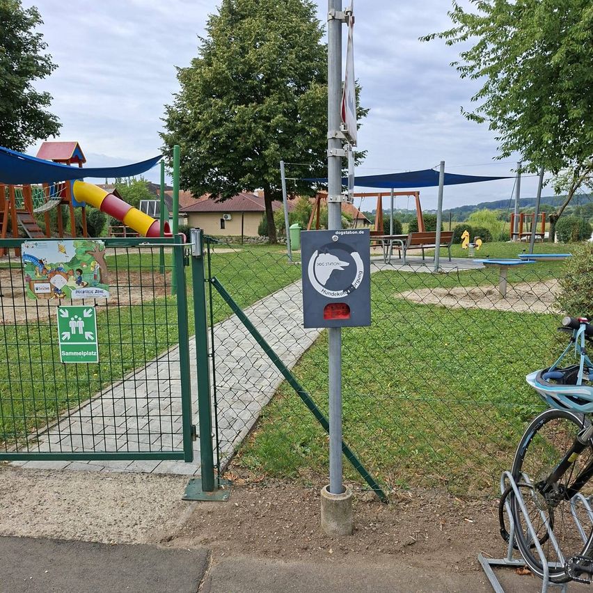 Ein Spielplatzbereich mit einer Hundestation und einem geschlossenen Tor. Fahrräder sind an der Seite geparkt. Bäume und Häuser im Hintergrund.