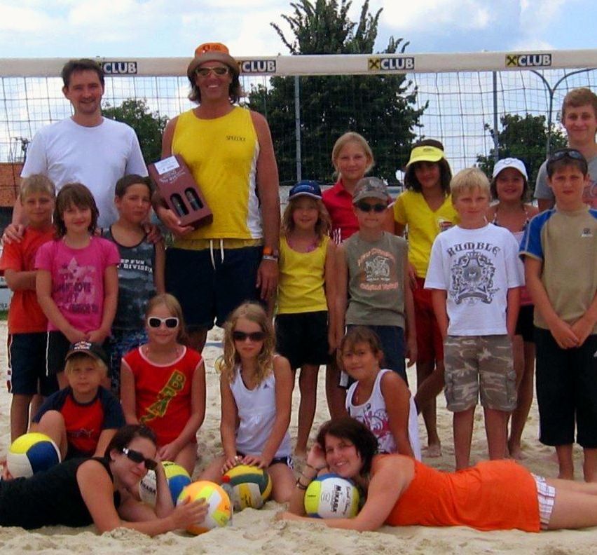 Eine Gruppe von Kindern und Erwachsenen posiert für ein Foto auf einem Beachvolleyballfeld. Ein Mann hält einen Pokal. Die Kinder tragen verschiedene farbige T-Shirts und Shorts. Im Hintergrund befindet sich ein Volleyballnetz.